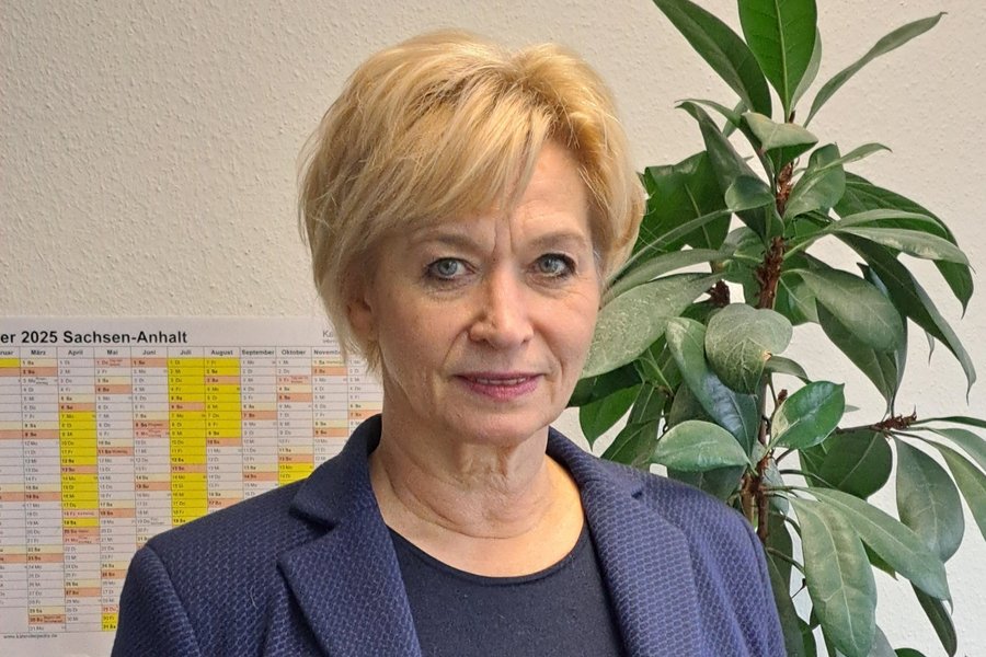 Foto vone einer Frau mit blonden Haaren und lilafarbenem Jacket - sie steht vor einer Wand mit einem Kalender und einer Pflanze