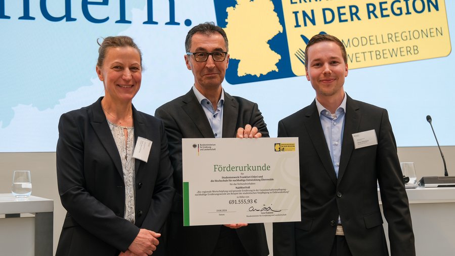 3 Personen nebeneinander, links eine Frau, in der Mitte Cem Özdemir, der eine Förderurkune in den Händen hält und rechts ein Mann
