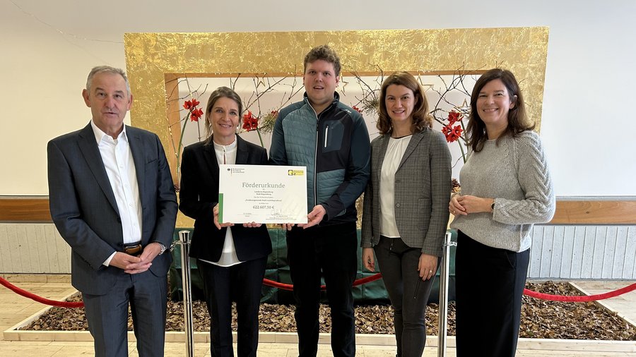5 Personen stehen nebeinander vor einem großen Bild, die zweite und dritte Person von links (eine Frau, ein Mann) halten ein Schild mit dem Schriftzug Förderurkunde hoch