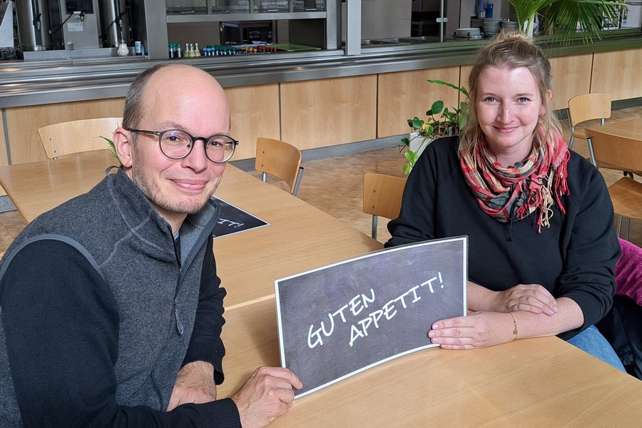Ein Mann und eine Frau sitzen sich an einem Holztisch gegenüber und schauen in die Kamera. Sie halten ein Schild mit der Aufschrift Guten Appetit zwischen ihren Händen